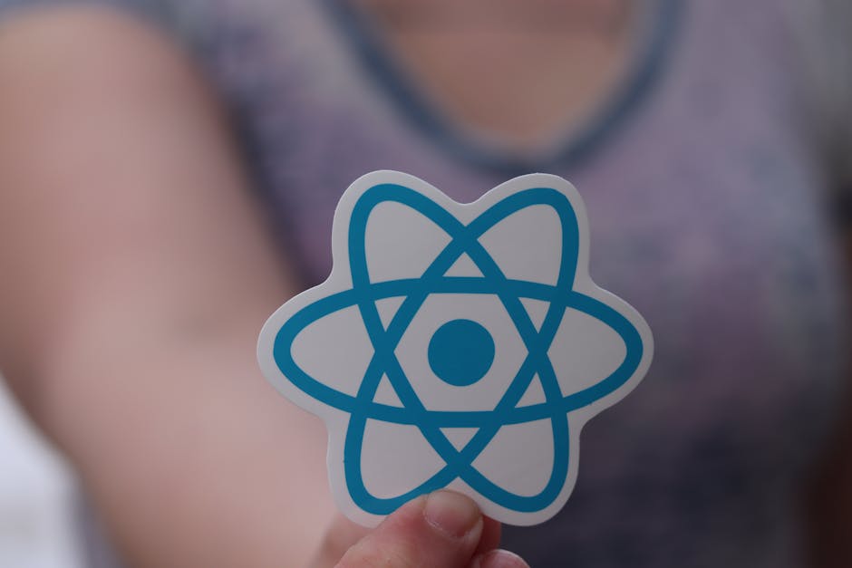 Close-up of a person holding a circular sticker with a blue atomic symbol on it, in front of a blurred background showing a person wearing a sleeveless top with a rounded neckline. The setting appears